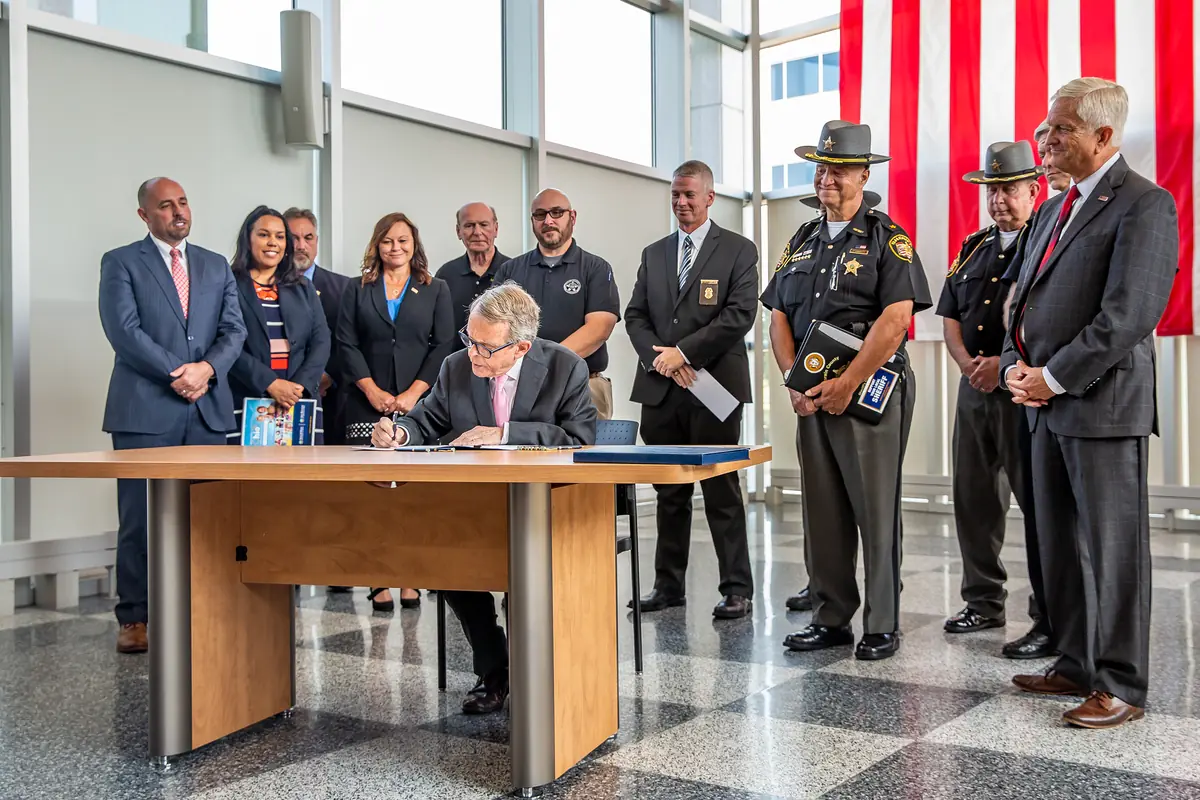Ohio Governor Mike DeWine seated at a desk signing an executive order, with Commander Lowe and law enforcement leaders standing behind him